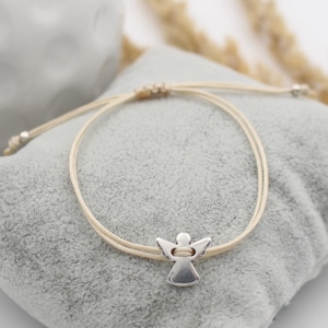 beiges Makramee Armband mit silberfarbenem Schutzengel auf grauem Kissen gelegen