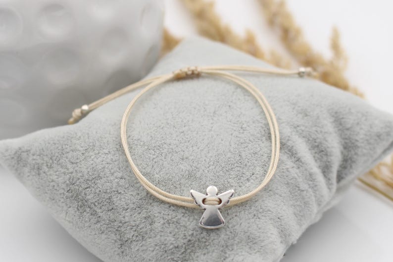 beiges Makramee Armband mit silberfarbenem Schutzengel auf grauem Kissen gelegen