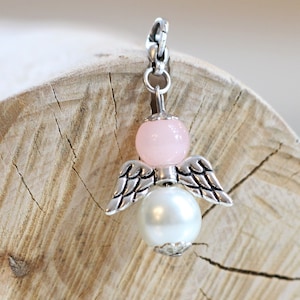 Könnte beinhalten: Ein silberner Engelsanhänger mit rosa und weißen Perlen. Der Engel hat Flügel und einen Heiligenschein. Der Anhänger hängt an einer silbernen Kette.