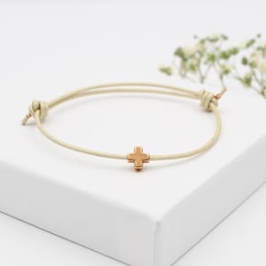 Könnte beinhalten: Ein Armband mit einer dünnen, hellbeigen Kordel, verstellbaren Schiebeknoten und einem kleinen, mittig platzierten metallischen Kreuzanhänger in warmem Roségold. Die Kordel ist glatt und kreisförmig und bietet ein schlichtes Design.