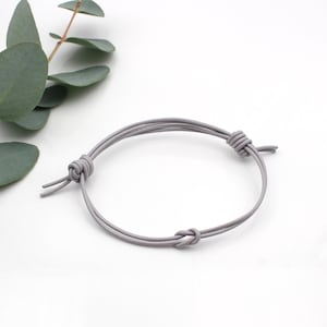 Könnte beinhalten: Ein hellgraues Kordelarmband mit glatter, abgerundeter Textur. Das Design besteht aus zwei parallelen Kordelsträngen, die mit drei markanten Knoten gesichert sind und eine verstellbare Schlaufenstruktur bilden, die eine kreisförmige Form beibehält.