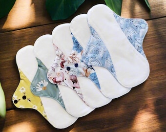May include: Five reusable cloth menstrual pads in various patterns. The pads are white with different colored patterns on the top. The patterns include a blue and white rainbow design, a floral design, and a green and yellow daisy design.