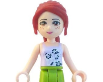 Puede incluir: Una muñeca de pelo rojo con una camisa blanca con estampado floral negro y pantalones verdes con diseños de mariposas moradas. La muñeca lleva sandalias moradas.