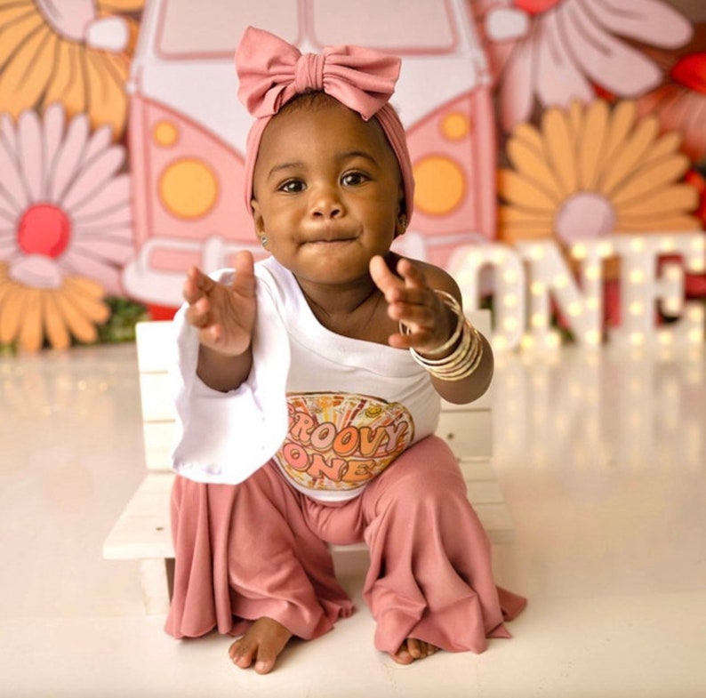 May include: A baby girl wearing a white shirt with the text "Groovy One" in a retro font and pink bell bottom pants. She is wearing a pink bow headband and has a light brown complexion. She is sitting on a white bench in front of a colorful backdrop with a flower design.