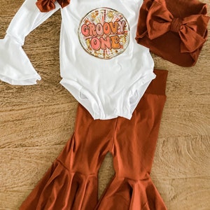 May include: A white baby onesie with the text "Groovy One" in a colorful retro font. The onesie is paired with brown bell bottom pants and a brown headband with a bow.