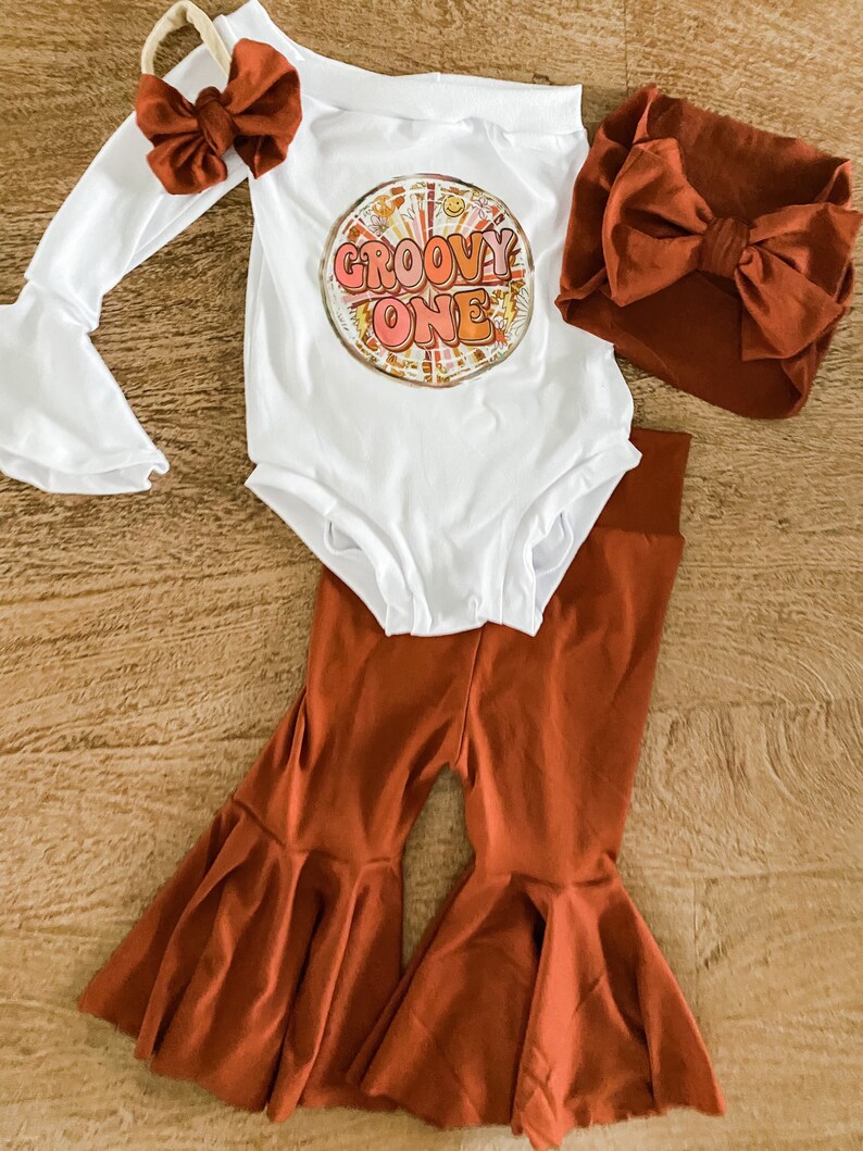 May include: A white baby onesie with the text "Groovy One" in a colorful retro font. The onesie is paired with brown bell bottom pants and a brown headband with a bow.