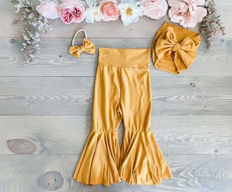May include: A mustard yellow baby outfit with a matching headband and a bow. The outfit includes a pair of flared pants and a headband with a bow.