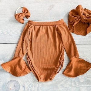 May include: A rust-colored ribbed bodysuit with long sleeves and bell bottoms. The bodysuit has an off-the-shoulder neckline. The outfit is paired with a matching rust-colored headband and a matching rust-colored turban.