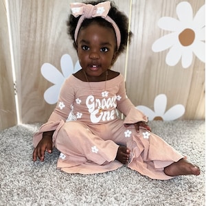 May include: A baby girl wearing a pink outfit with white flowers and the text "Groovy One" on the front. She is wearing a pink headband with a white flower on it. The baby is sitting on a light brown rug.