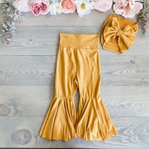 May include: Mustard yellow flared pants with a matching mustard yellow bow headband. The pants have a wide leg and a ruffled hem.