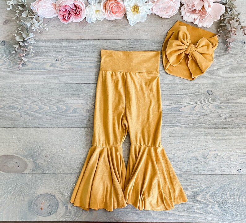 May include: Mustard yellow flared pants with a matching mustard yellow bow headband. The pants have a wide leg and a ruffled hem.