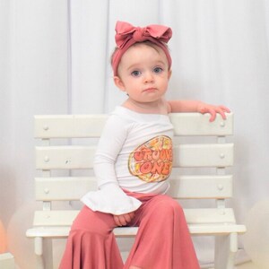 May include: A baby girl wearing a white long-sleeve shirt with a peach-colored graphic that says "Groove One" and peach-colored bell bottom pants. She is wearing a pink headband with a large bow. She is sitting on a white wooden bench.