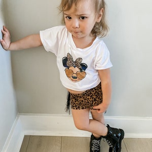 May include: A young girl wearing a white t-shirt with a cartoon character graphic and leopard print shorts. She is wearing black boots and has her hair pulled back in a ponytail.