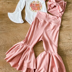 May include: A baby outfit set with a white long-sleeve bodysuit with the text "Groovy One" in a retro font, a pink headband, a pink bow, and a pair of pink bell-bottom pants.