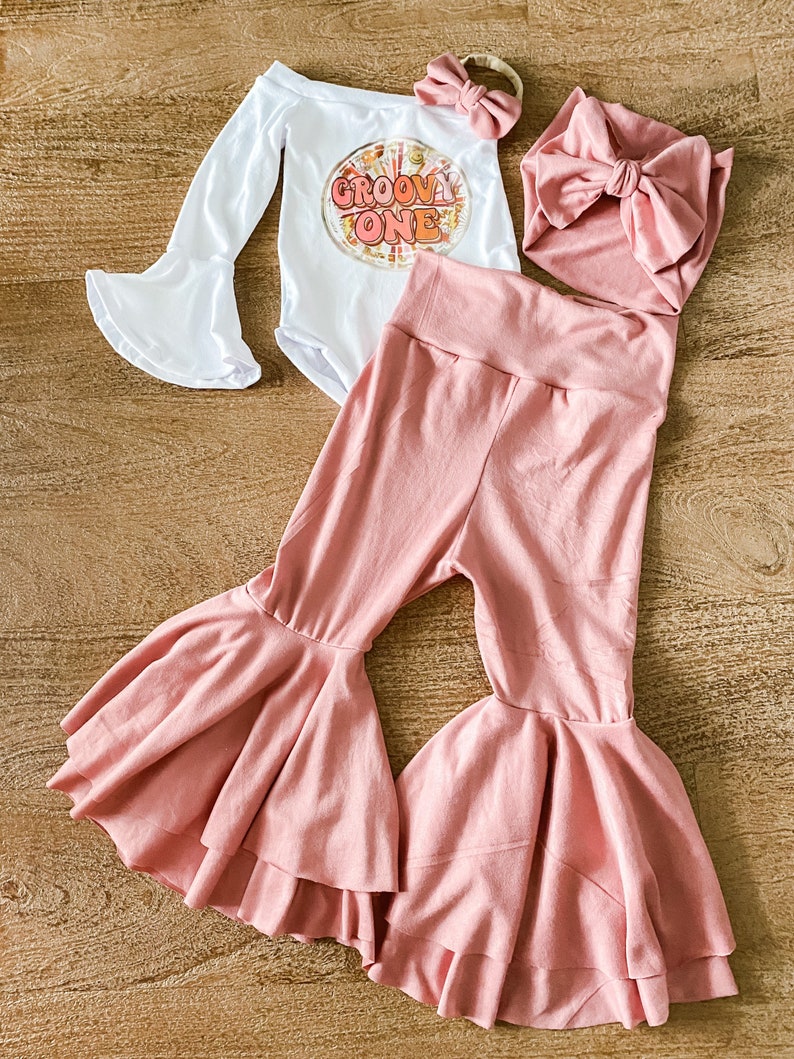 May include: A baby outfit set with a white long-sleeve bodysuit with the text "Groovy One" in a retro font, a pink headband, a pink bow, and a pair of pink bell-bottom pants.