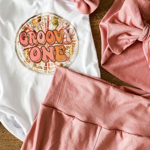 May include: A white onesie with the text "Groovy One" in a retro font and a colorful graphic design. The onesie is paired with a pink headband and a pink knotted headband.