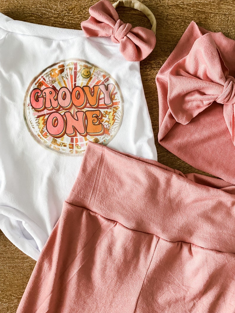 May include: A white onesie with the text "Groovy One" in a retro font and a colorful graphic design. The onesie is paired with a pink headband and a pink knotted headband.