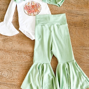 May include: A baby outfit set with a white long-sleeved bodysuit with a groovy one graphic, a green headband, and green bell bottom pants.