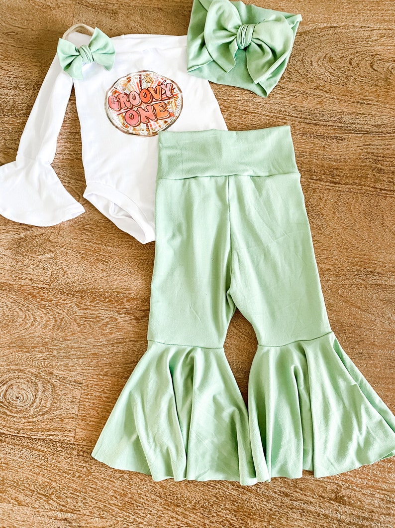 May include: A baby outfit set with a white long-sleeved bodysuit with a groovy one graphic, a green headband, and green bell bottom pants.