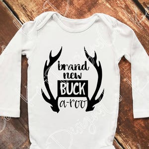 May include: White long-sleeve baby onesie with a black graphic of a deer antler with the text "brand new buck a-roo".