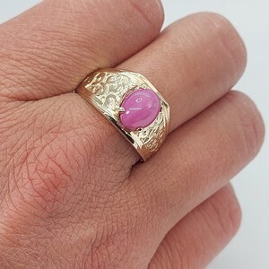 Op de afbeelding: Een gouden ring met een roze ovale edelsteen, gezet in een gedetailleerd, versierd ontwerp.