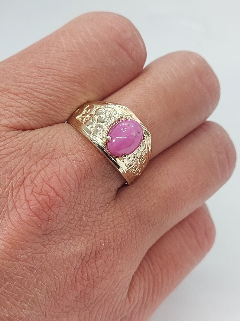Op de afbeelding: Een gouden ring met een roze ovale edelsteen, gezet in een gedetailleerd, versierd ontwerp.
