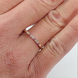 Pode incluir: Um delicado anel de banda de ouro com uma fileira de pequenas pedras preciosas rosa e brancas.