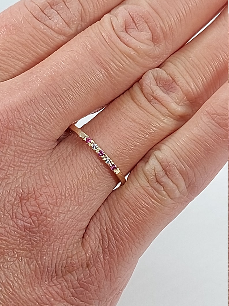 Pode incluir: Um delicado anel de banda de ouro com uma fileira de pequenas pedras preciosas rosa e brancas.