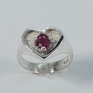 May include: A silver ring with a heart-shaped design. The heart is filled with a textured pattern and features a red gemstone in the centre.