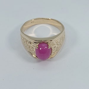 Op de afbeelding: Een gouden ring met een ovale roze edelsteen in het midden. De ring heeft een gedetailleerd ontwerp rond de edelsteen.