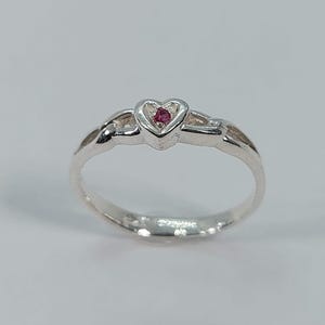 May include: A silver ring with a heart-shaped setting. The heart holds a small, round, red gemstone. The ring has a delicate, intertwined design on the band, and is set against a plain background.