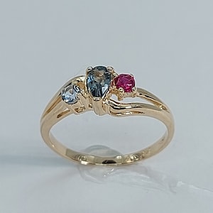 Peut inclure: Une bague en or sertie de trois pierres précieuses. La plus grande pierre est une pierre bleue en forme de poire. Les deux autres pierres sont rondes, l'une est rouge et l'autre est bleue.