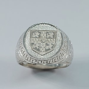 May include: A silver ring with a detailed design of a coat of arms on the top. The ring has a textured band with a Greek key pattern.