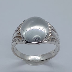 May include: A silver signet ring with a smooth, rounded top and an intricate, engraved design around the band.