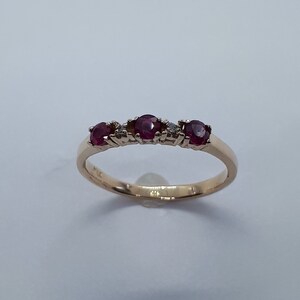 May include: A gold band ring with five red gemstones and two small diamonds. The ring is set on a white background.