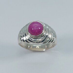 K&ouml;nnte beinhalten: Ein silberner Ring mit einem rosafarbenen sternf&ouml;rmigen Edelstein, der in einer erh&ouml;hten Fassung sitzt. Die Band ist mit einem wirbelnden Muster texturiert.