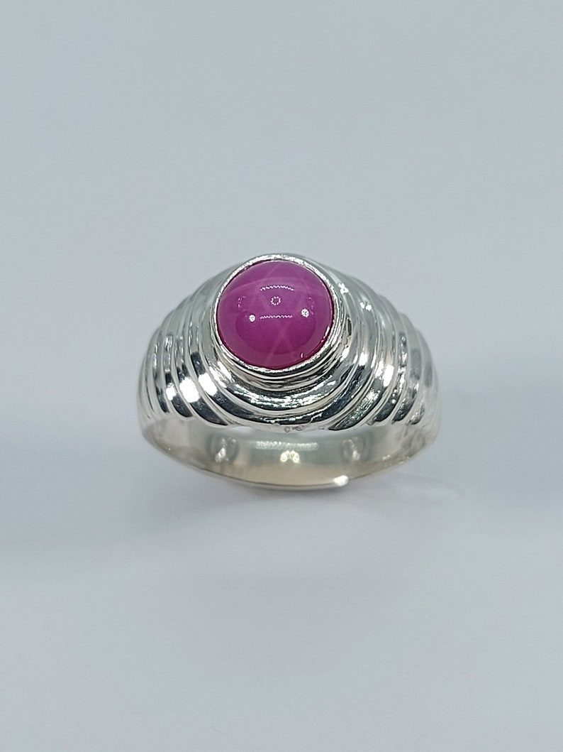 K&ouml;nnte beinhalten: Ein silberner Ring mit einem rosafarbenen sternf&ouml;rmigen Edelstein, der in einer erh&ouml;hten Fassung sitzt. Die Band ist mit einem wirbelnden Muster texturiert.