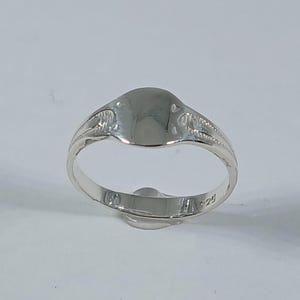 May include: A polished silver signet ring with an oval face and decorative detailing on the shoulders. The ring is set against a white background, showcasing its reflective surface. The ring is a classic piece of jewellery.