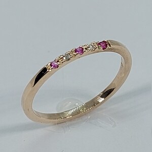 Pode incluir: Um delicado anel de banda de ouro com uma fileira de pequenas pedras preciosas rosa e brancas.