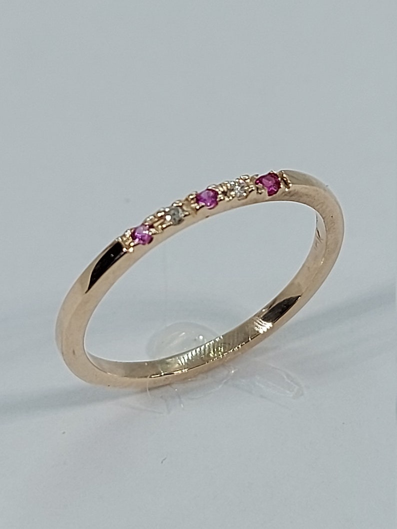 Pode incluir: Um delicado anel de banda de ouro com uma fileira de pequenas pedras preciosas rosa e brancas.