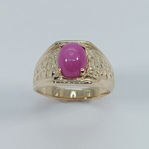 Op de afbeelding: Een gouden ring met een ovale roze edelsteen in het midden. De ringband is getextureerd met een decoratief ontwerp.