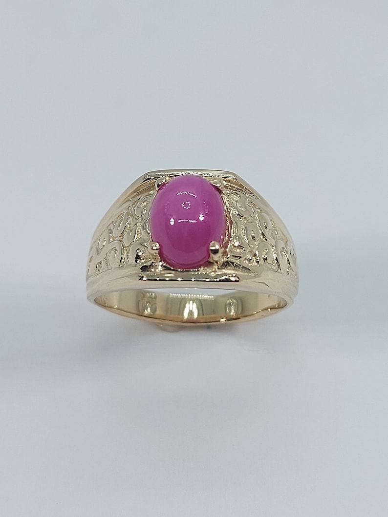 Op de afbeelding: Een gouden ring met een ovale roze edelsteen in het midden. De ringband is getextureerd met een decoratief ontwerp.