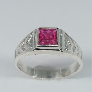 May include: A silver ring with a square-shaped pink gemstone set in a raised bezel. The ring has a detailed design on the band.