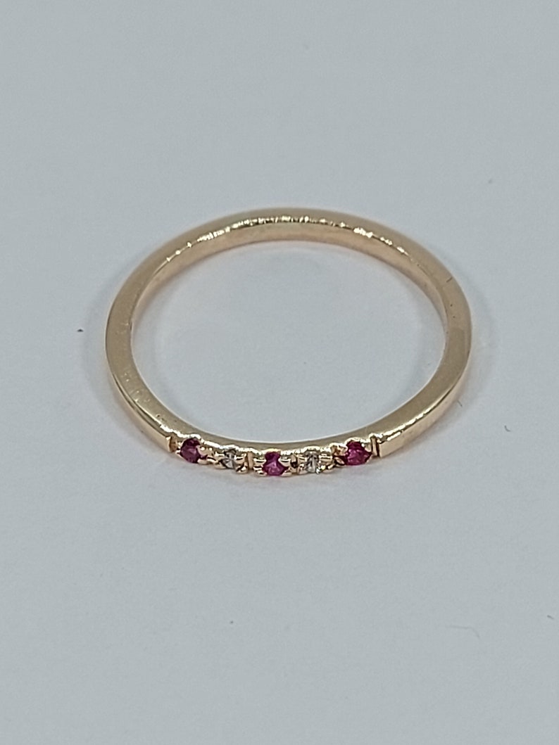 Pode incluir: Um delicado anel de banda de ouro com uma fileira de pequenas pedras preciosas vermelhas e brancas.