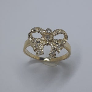 May include: A gold ring with a bow-shaped design, embellished with small, clear gemstones. The ring is set against a plain, light background. The jewellery piece is a fashion accessory. The ring's design is intricate and elegant.