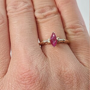 May include: A gold ring featuring a marquise-shaped ruby gemstone with two smaller baguette-shaped diamonds on either side.