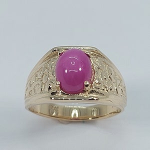 Op de afbeelding: Een gouden ring met een ovale roze edelsteen gezet in een gedetailleerd, versierd ontwerp.