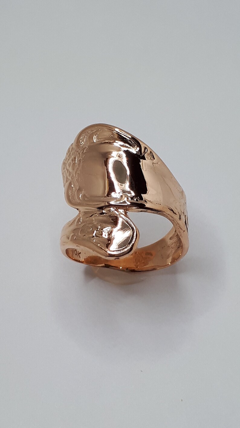 May include: A gold-toned ring made from a bent spoon. The ring is decorated with a floral design and has a shiny finish.