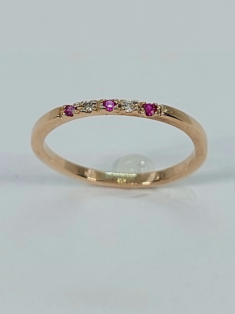 Pode incluir: Um delicado anel de banda de ouro com pedras preciosas rosa e brancas alternadas. O anel &eacute; engastado com pequenas pedras redondas.