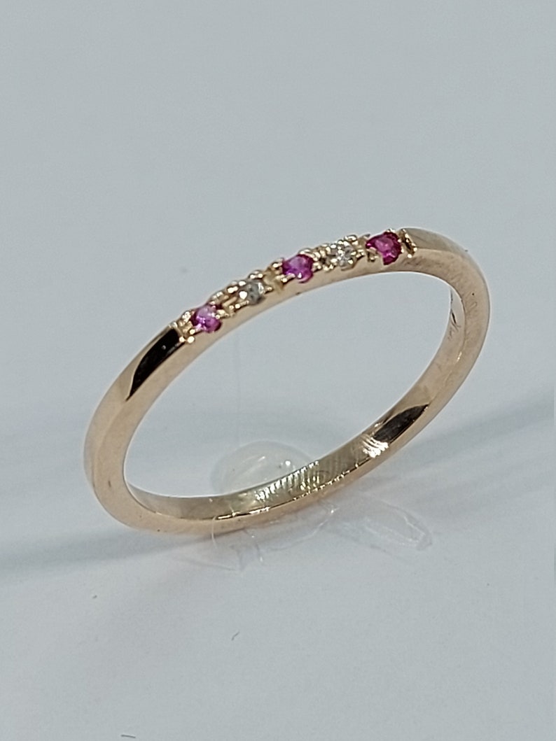 Pode incluir: Um delicado anel de banda de ouro com uma fileira de pequenas pedras preciosas rosa e brancas.